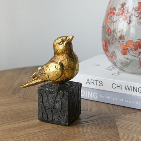 Gold bird figurine on a black base with a vase and books in the background
