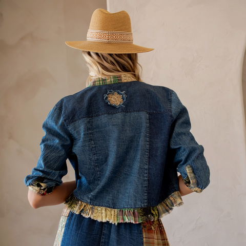 Person wearing a denim jacket and plaid dress with a straw hat, standing against a beige wall.