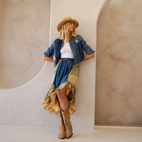 Woman in a stylish outfit with a denim jacket, patterned skirt, and cowboy boots against a neutral background.