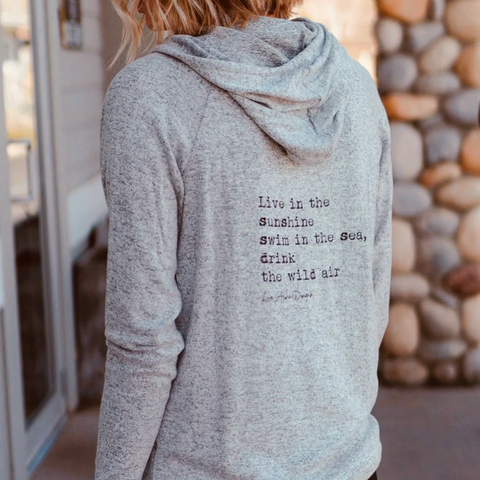 Person wearing a gray hoodie with a quote on the back, standing in front of a stone wall.