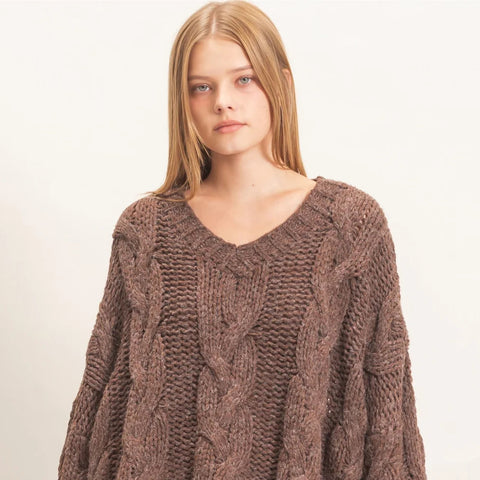 A woman wearing a chunky brown cable-knit sweater with a V-neck and black pants, photographed against a plain light background.
